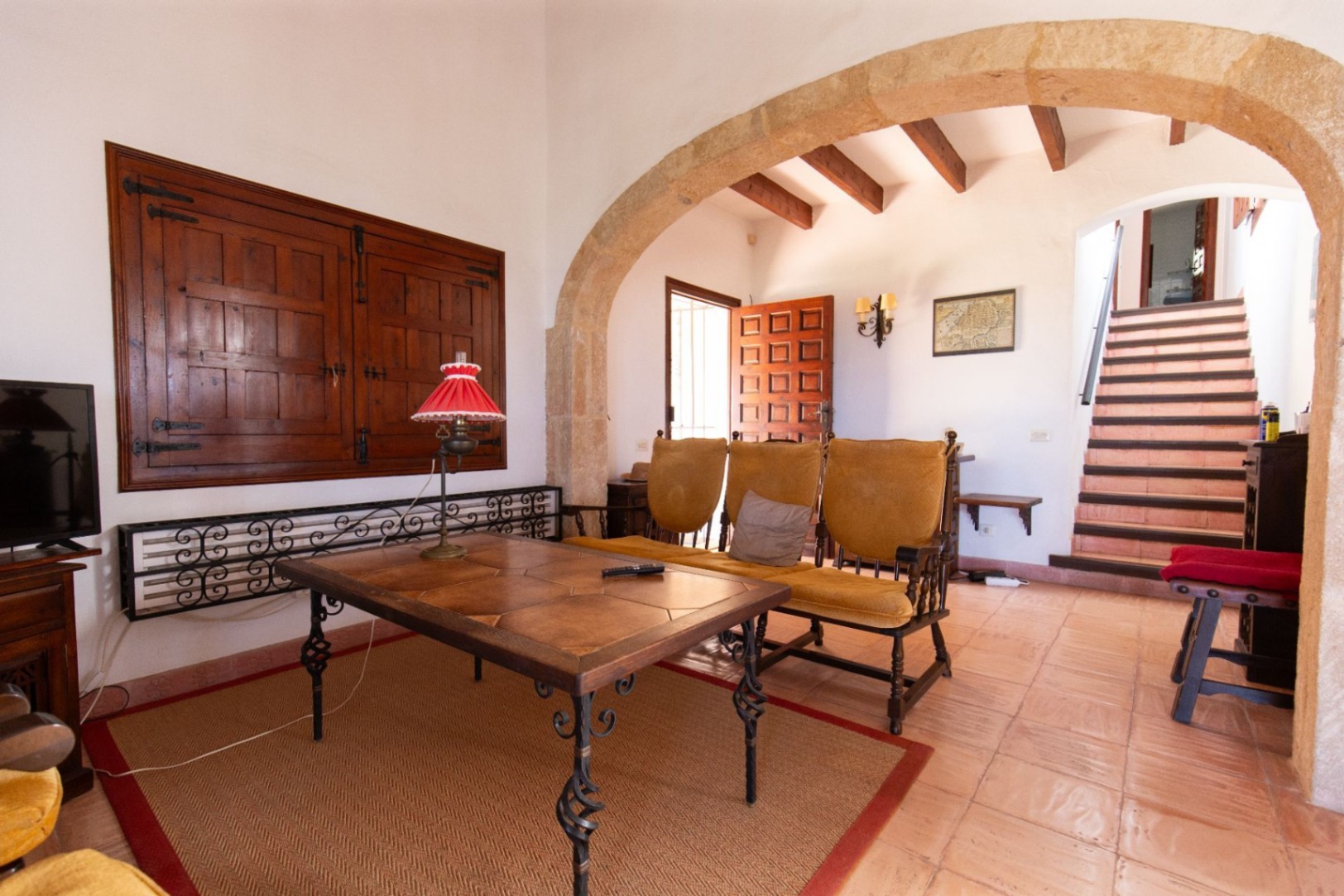 Living area featuring a stone archway, terracotta floors, and exposed wooden beams. Includes a rustic tiled table and stairs leading to the upper level.