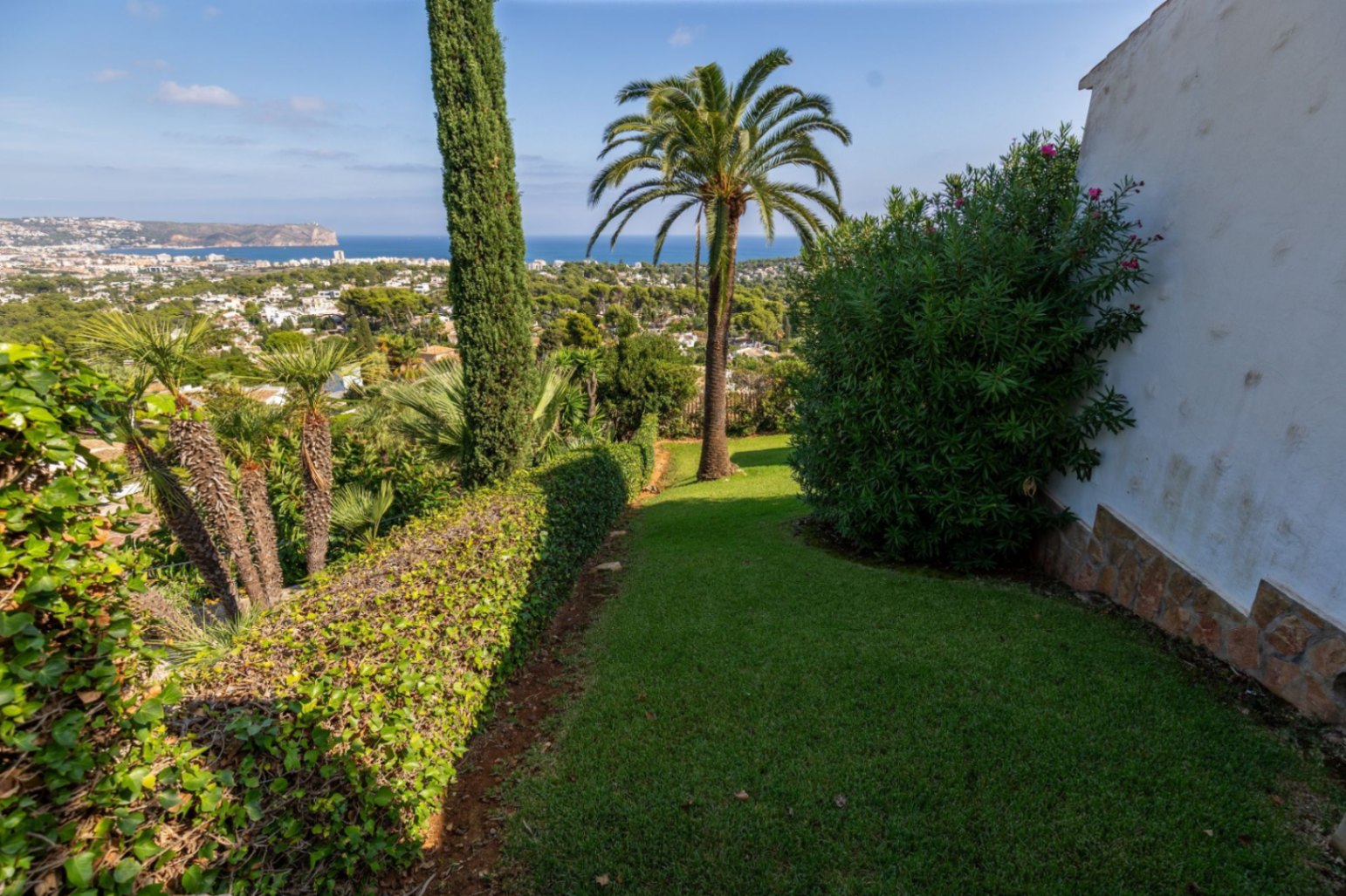 Manicured garden with a green lawn, palm trees, and cypress, offering panoramic views of the coastal town and the Mediterranean Sea.