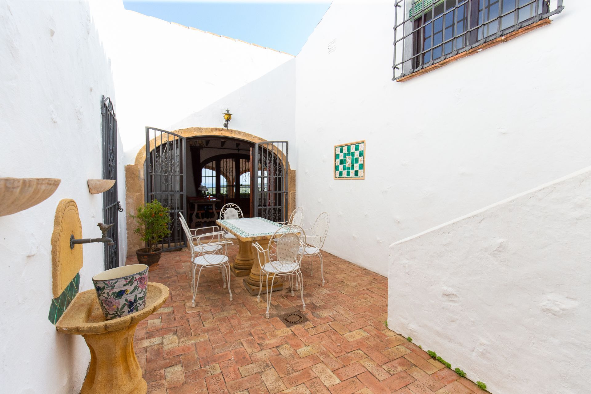 Mediterranean patio featuring patterned terracotta tiles, a stone fountain, and white wrought iron seating. An arched entrance with decorative metal gates leads inside.