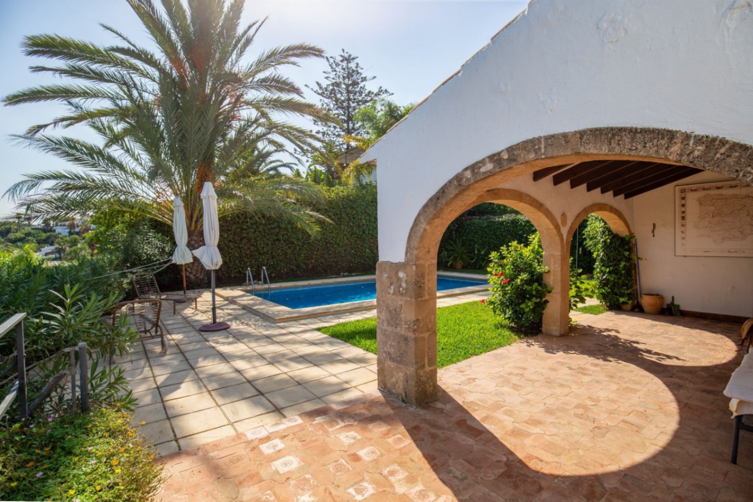 Porch with stone arches and terracotta flooring overlooking a private swimming pool, surrounded by palm trees and mature Mediterranean gardens.