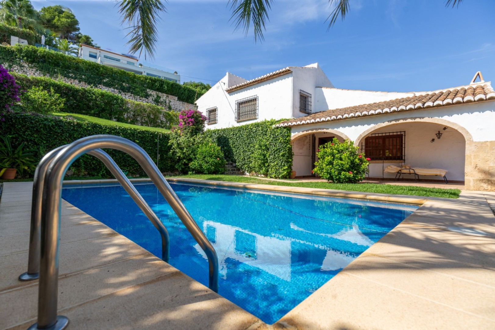 Rectangular swimming pool with blue mosaic tiles and stainless steel ladder, adjacent to a traditional white-washed villa with arched porches and terracotta roof.