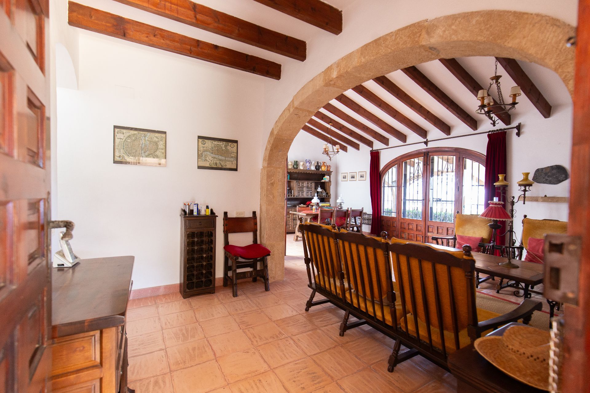 Rustic living area featuring exposed dark wood beams, a prominent stone archway, and terracotta flooring. Arched wooden doors lead to the exterior, providing natural light.