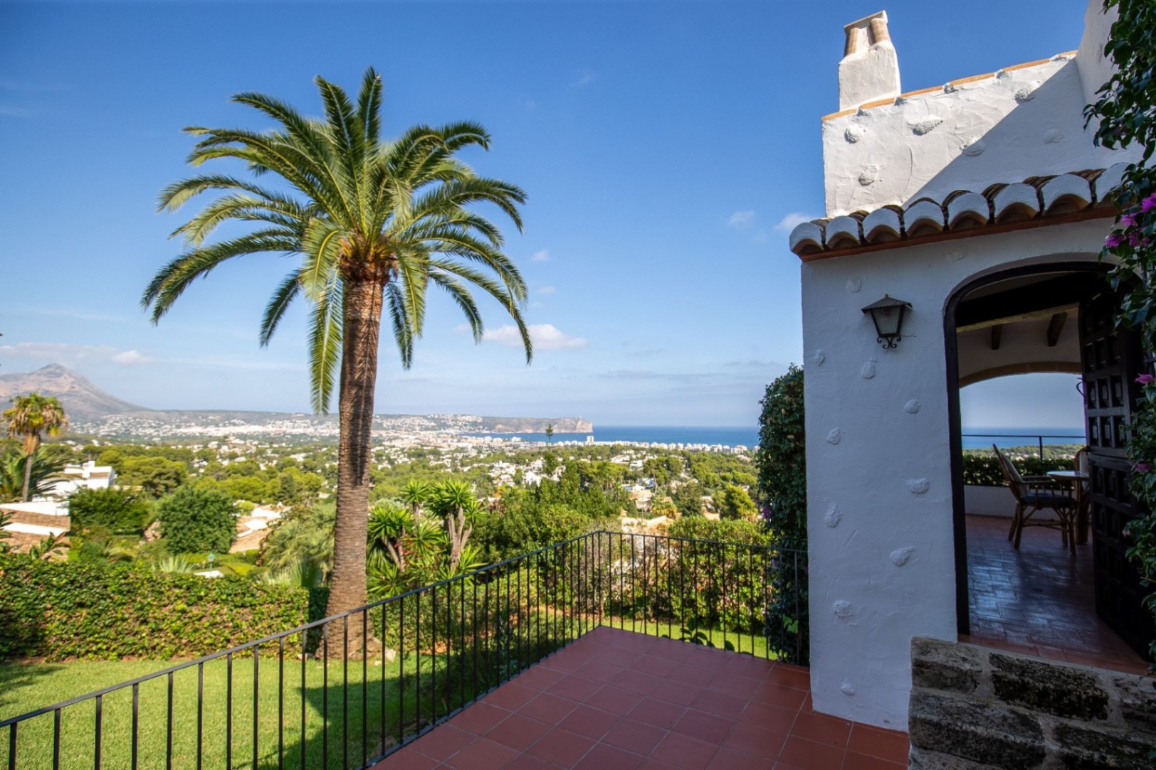 Terrace with terracotta tiles and iron railing overlooking the Mediterranean coastline, featuring a large palm tree and traditional white stucco architecture.