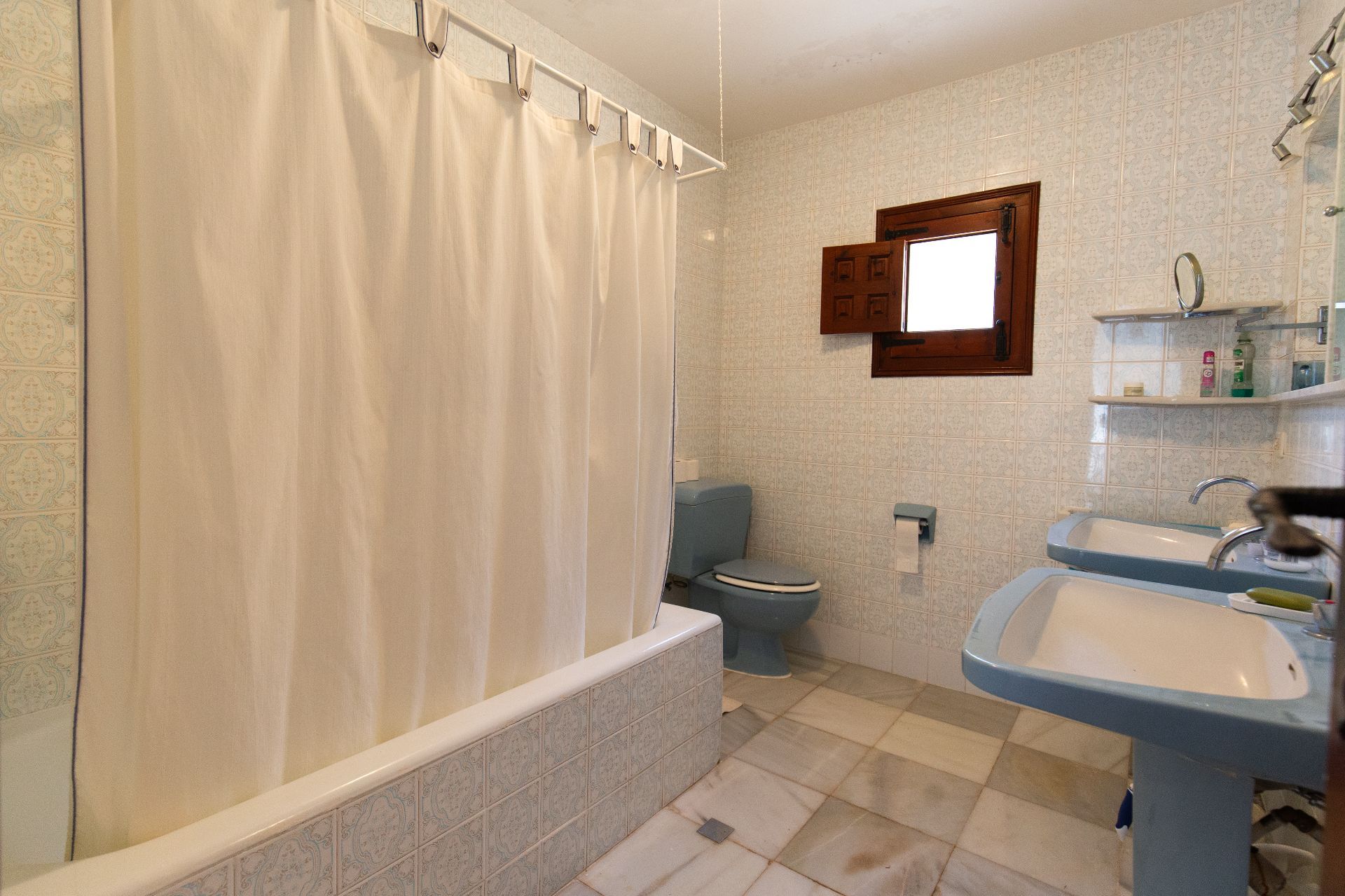 Traditional bathroom featuring double blue pedestal sinks, a matching toilet, and a tiled bathtub. The space includes patterned wall tiles and a small wooden window.