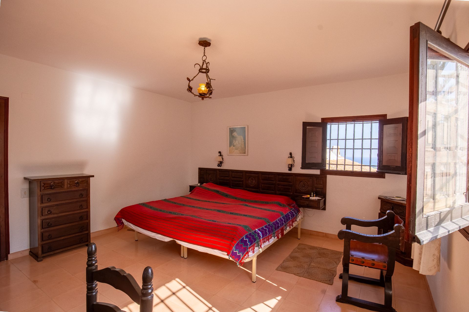 Traditional bedroom featuring a dark wood carved headboard, terracotta tile flooring, and a rustic wrought-iron chandelier. Natural light enters through a window with wooden shutters.