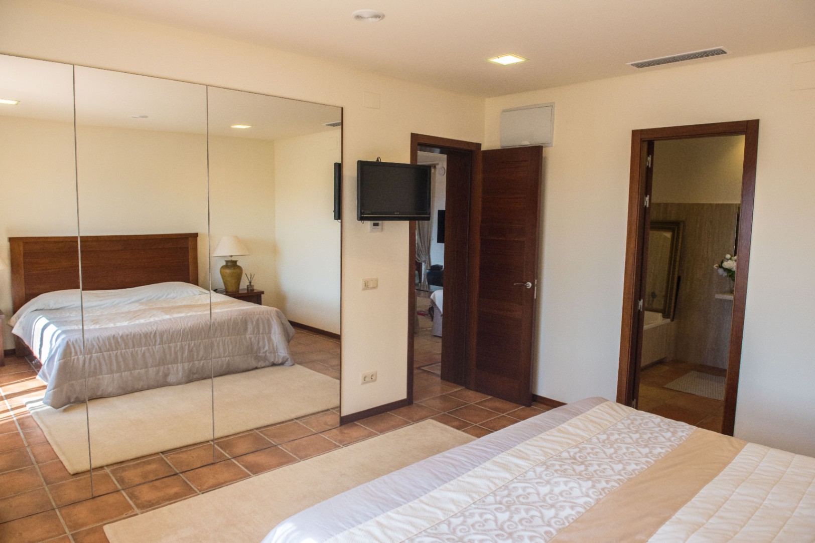 Bedroom with terracotta tile flooring, a large mirrored wardrobe, and a wall-mounted TV. Features include recessed lighting and direct access to an en-suite bathroom.
