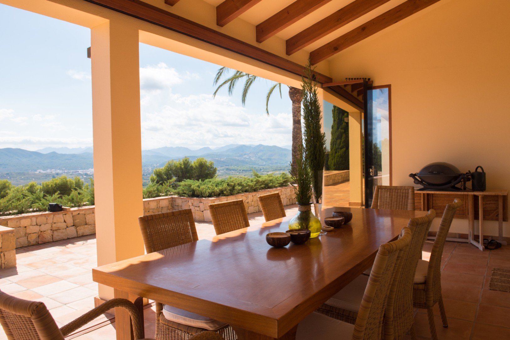 Covered terrace featuring a large wooden dining table and rattan chairs under exposed timber beams. The space offers panoramic mountain views and stone-walled borders.