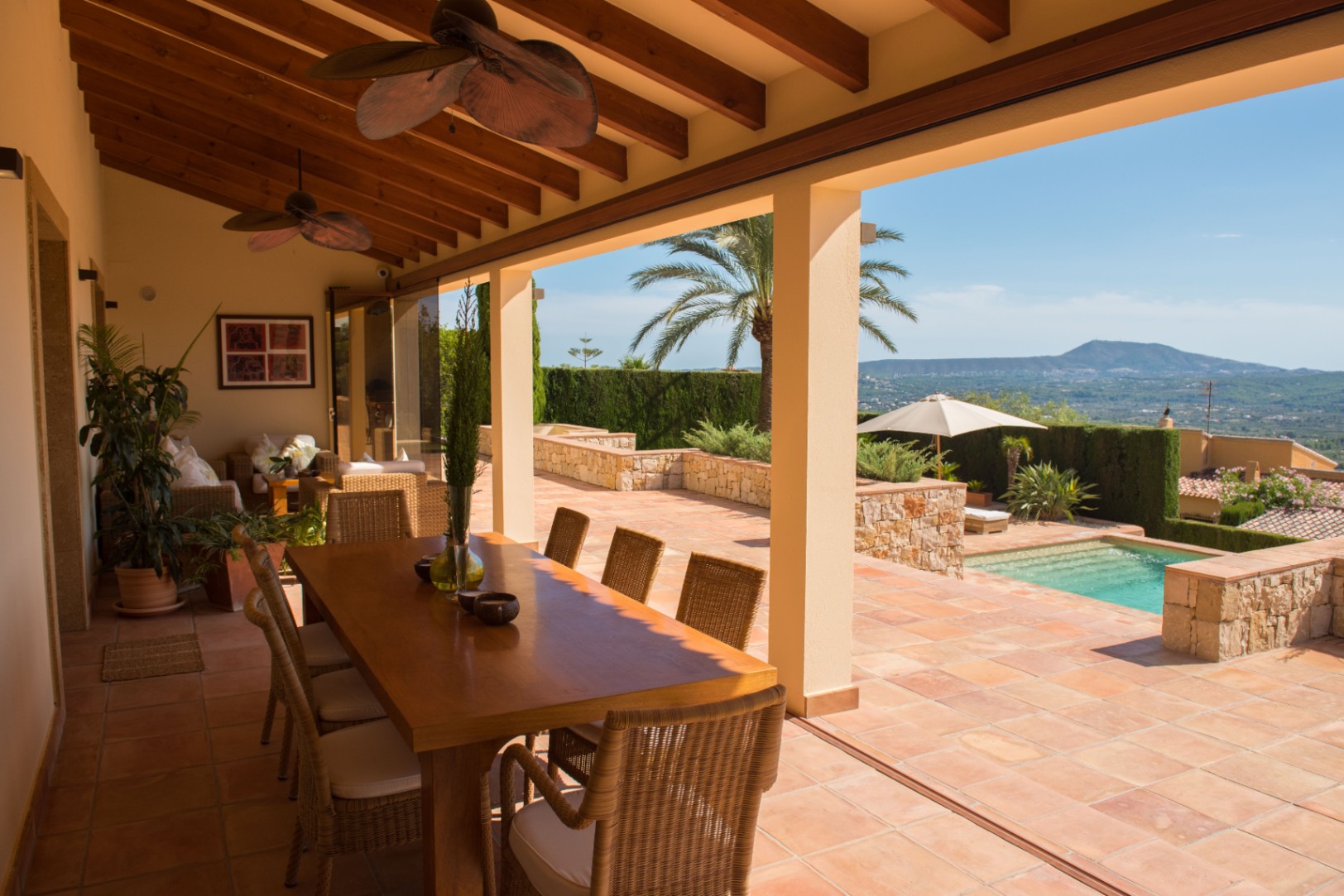 Covered terrace with exposed wooden beams, terracotta floors, and a dining area overlooking a private pool and distant mountain views.