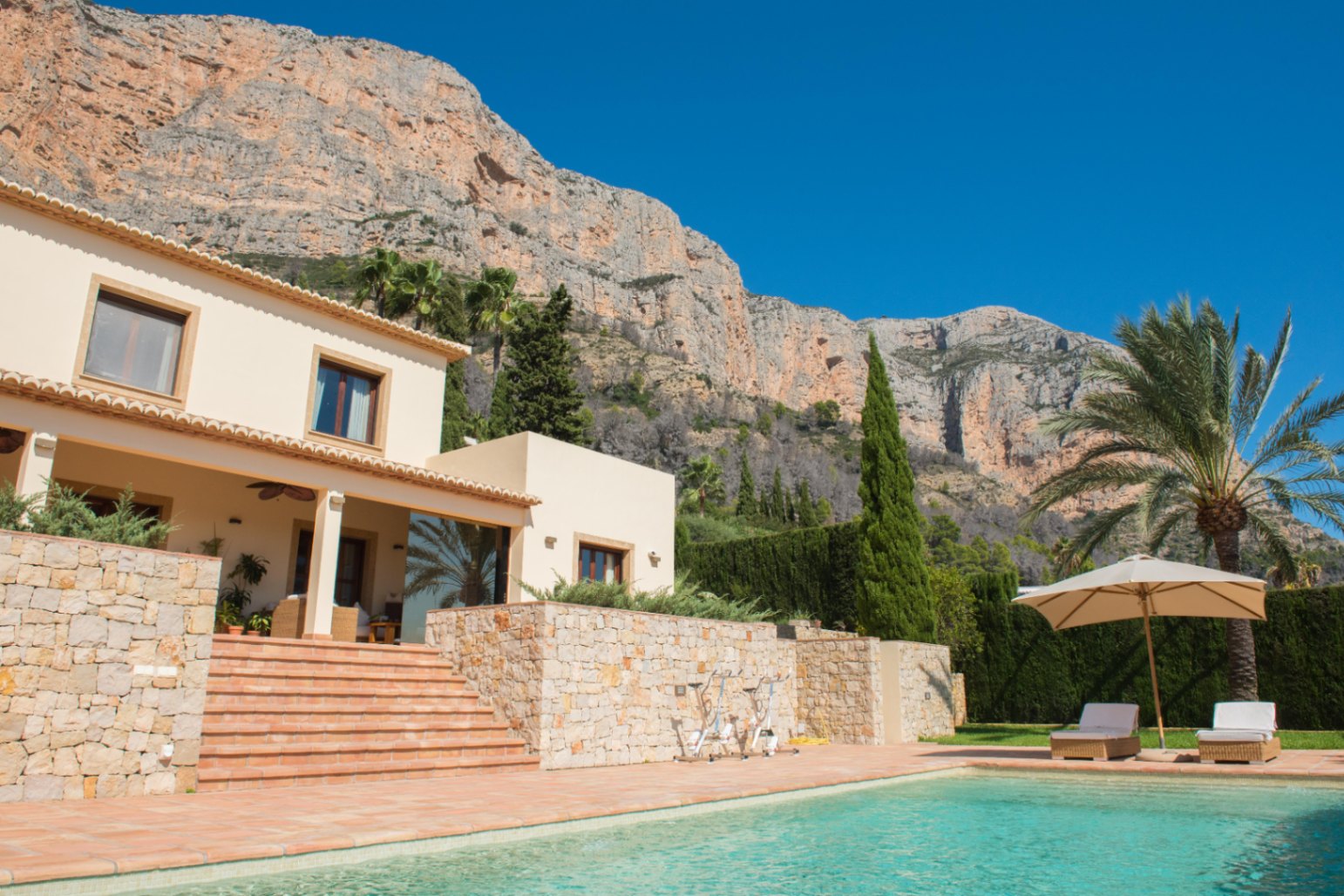 Exterior view featuring a turquoise pool, terracotta terrace, and stone walls set against a dramatic mountain backdrop with Mediterranean vegetation.