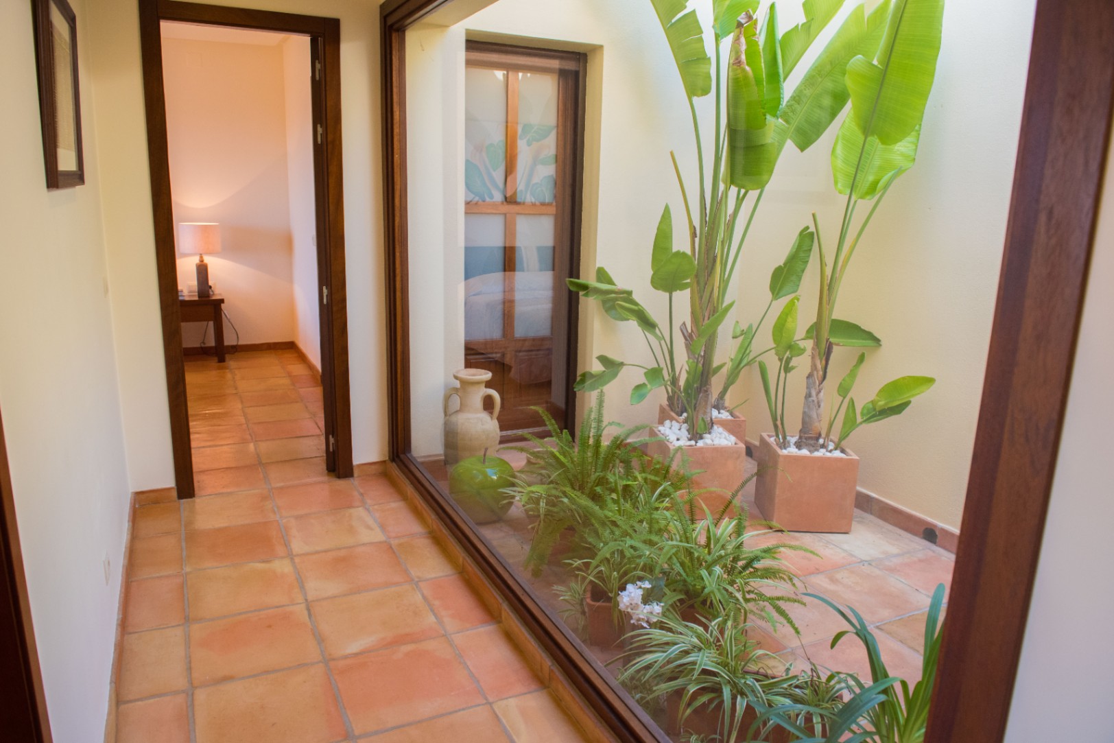 Hallway with terracotta flooring featuring a large glass window overlooking an internal patio with tropical plants and natural light.