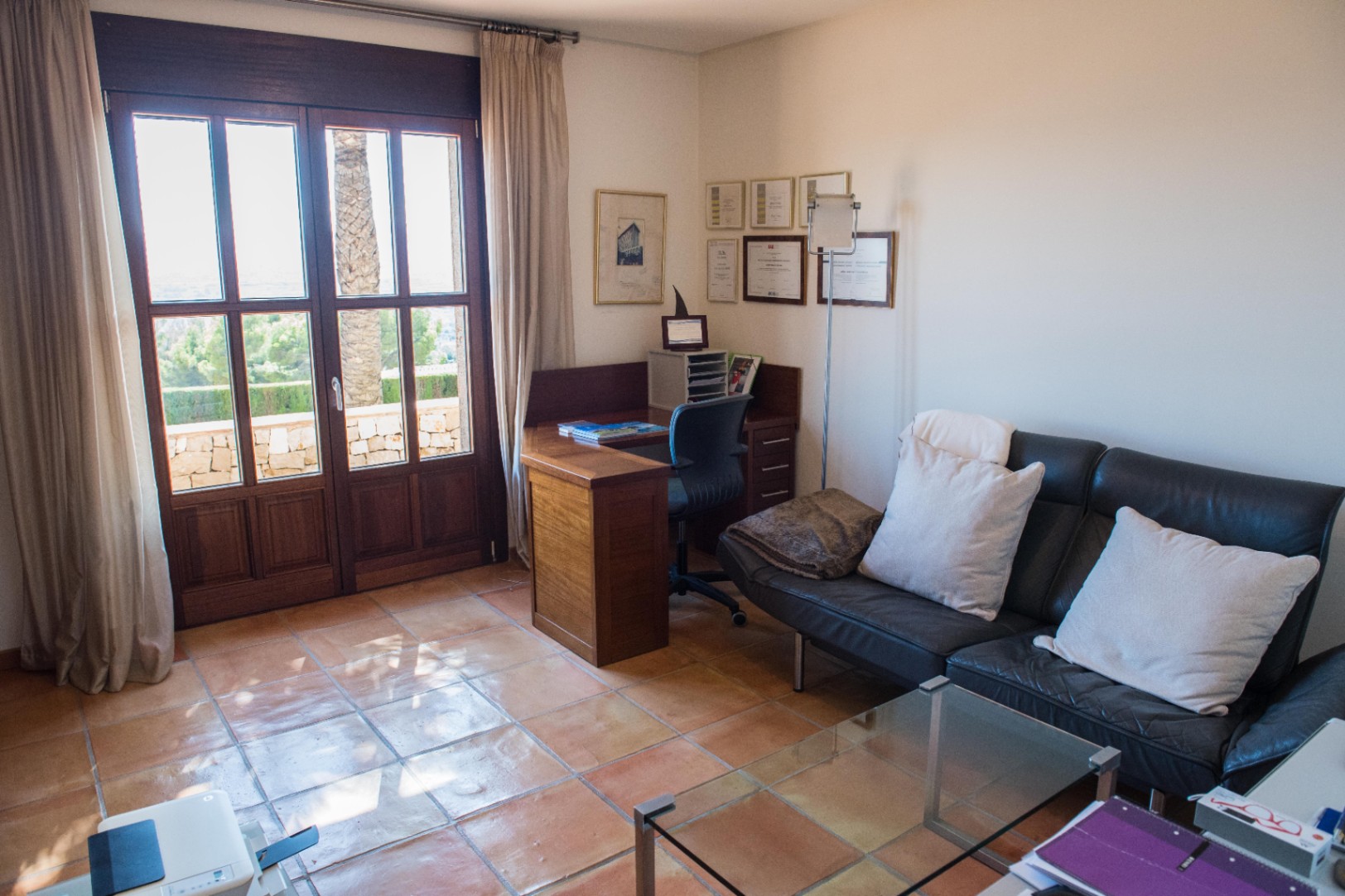 Home office with terracotta tile flooring, a wooden corner desk, and a black leather sofa. Large wooden glass doors provide natural light and access to a terrace.