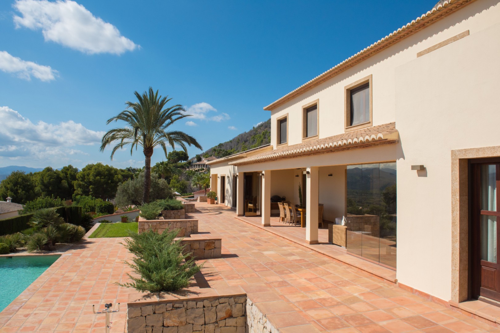 Mediterranean villa exterior with a terracotta terrace, stone-clad planters, and a covered porch. A palm tree overlooks the pool area and the surrounding mountain landscape.