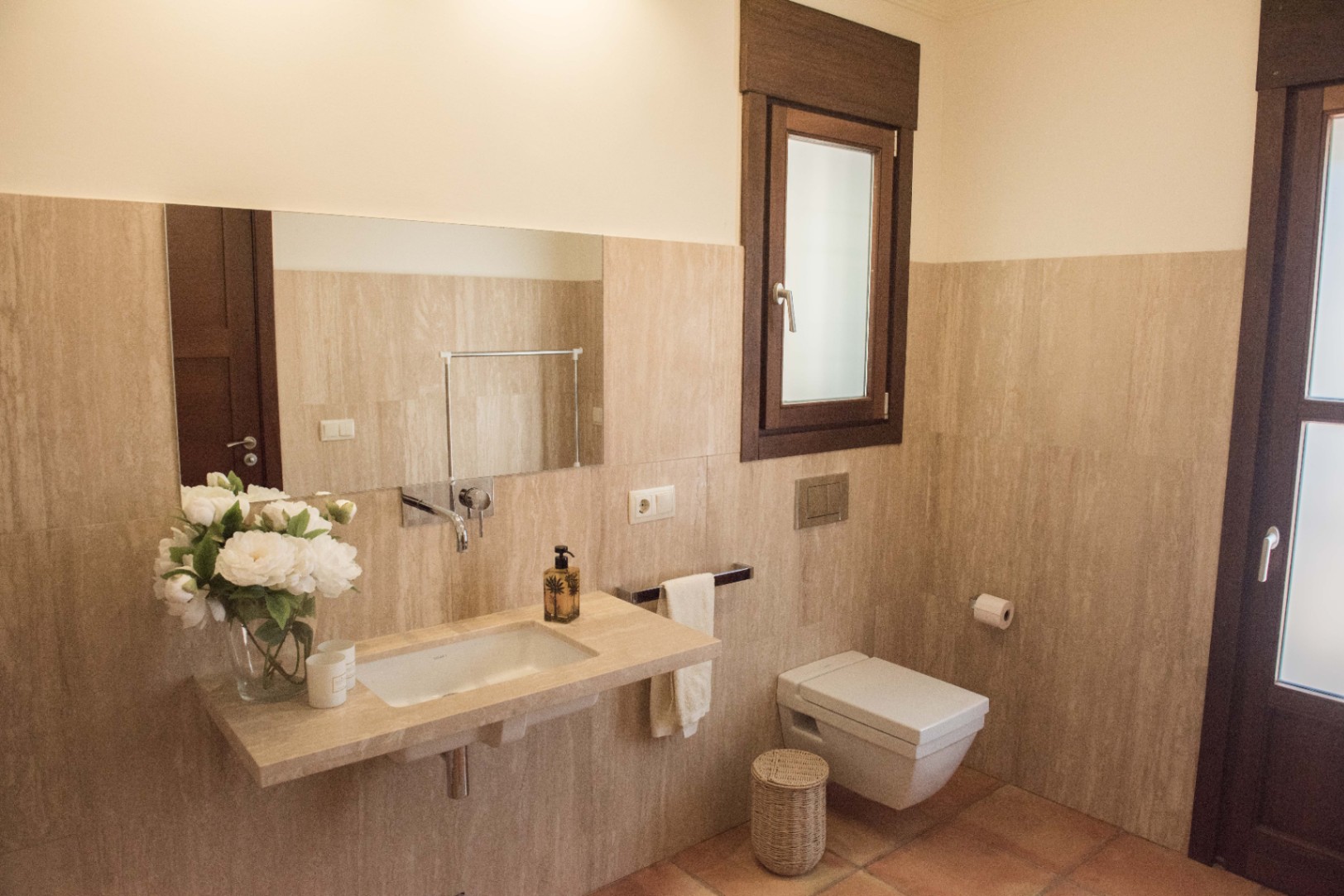 Modern bathroom featuring a floating stone vanity with integrated sink, wall-mounted chrome fixtures, and a wall-hung toilet against travertine walls.
