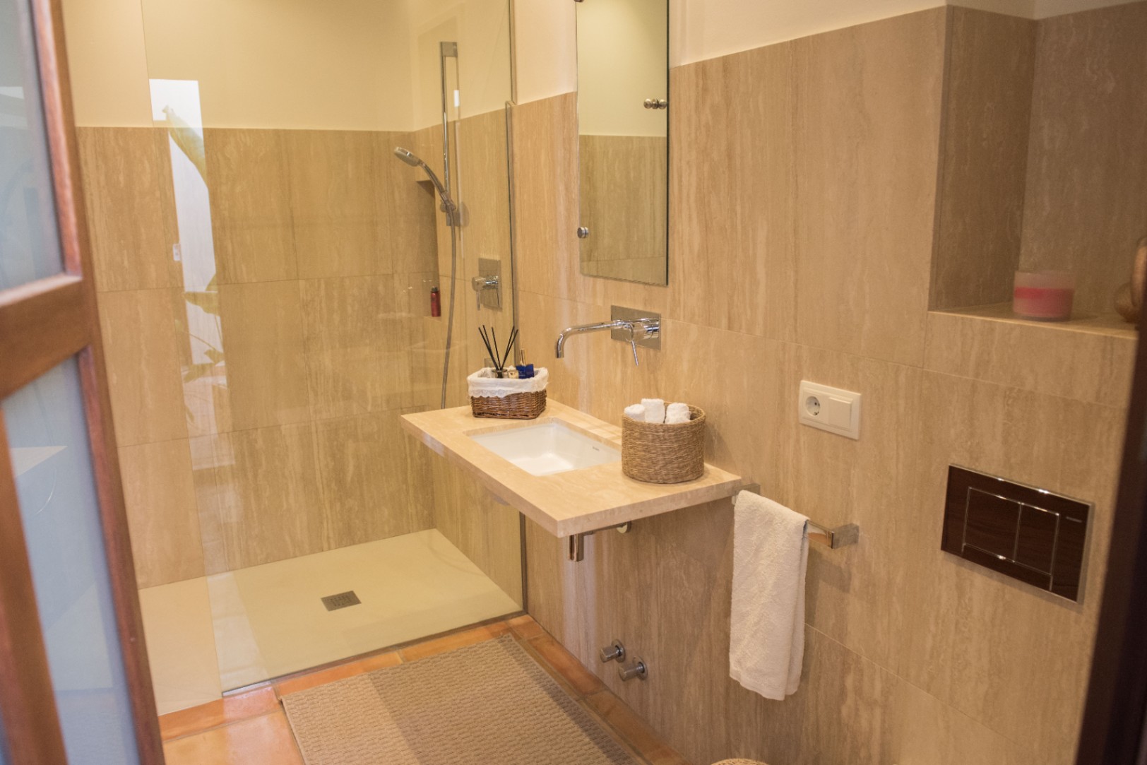 Modern bathroom featuring a walk-in shower with glass partition, floating stone vanity, and wall-mounted chrome fixtures against beige travertine tiling.