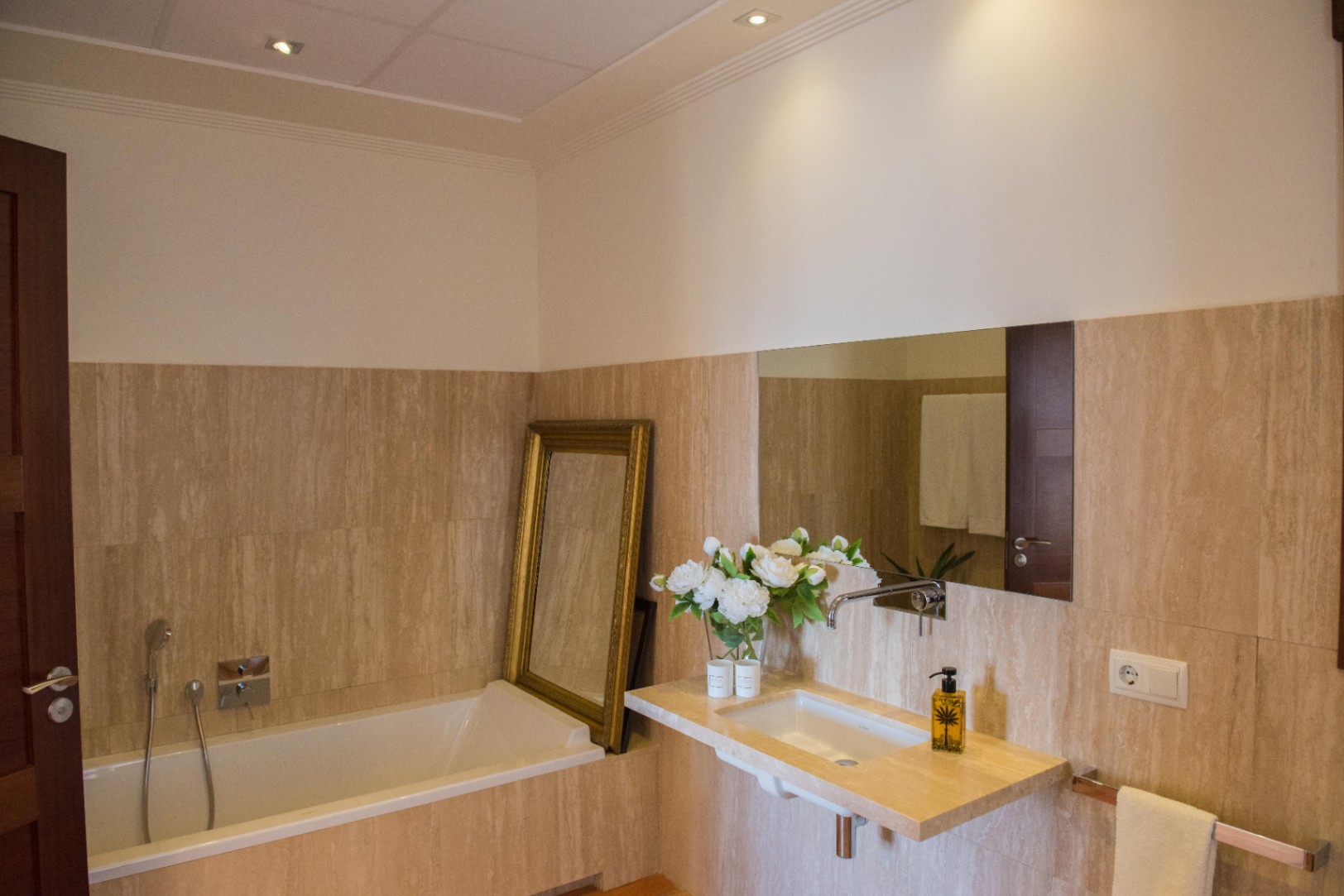 Modern bathroom featuring travertine stone walls, a floating vanity with integrated sink, and a built-in bathtub with a gold-framed mirror accent.