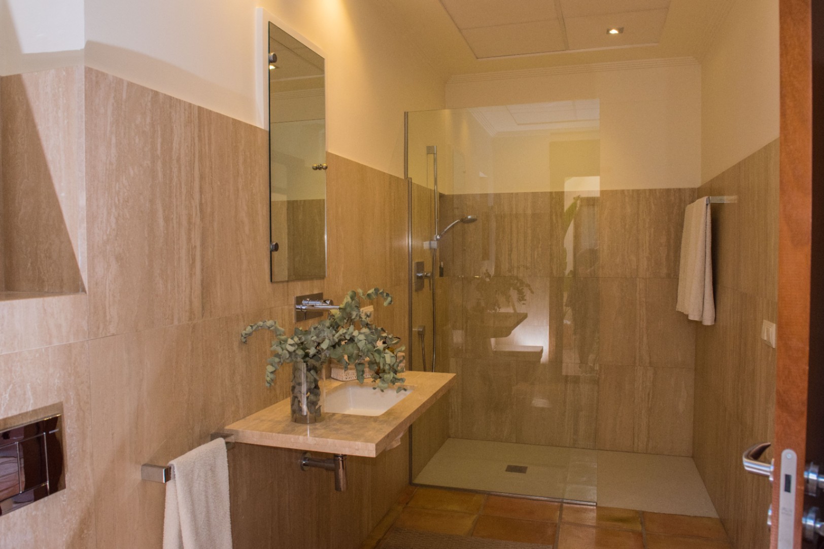 Modern bathroom featuring travertine wall tiling, a floating stone vanity, and a walk-in shower with a frameless glass partition and terracotta flooring.