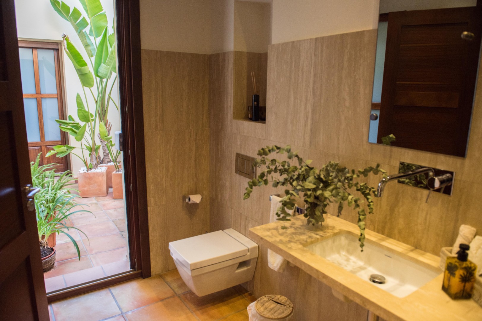 Modern bathroom featuring travertine wall tiling, a wall-mounted toilet, and an open door leading to a terracotta patio with lush tropical greenery.