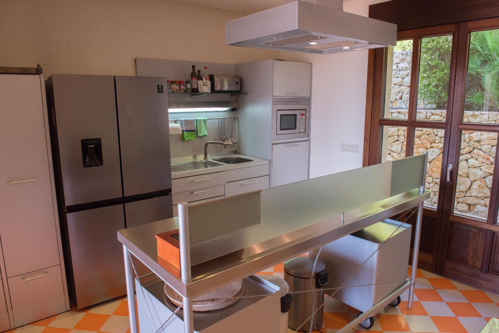 Modern kitchen featuring a stainless steel island, checkered tile flooring, and large wooden-framed glass doors overlooking a natural stone wall.