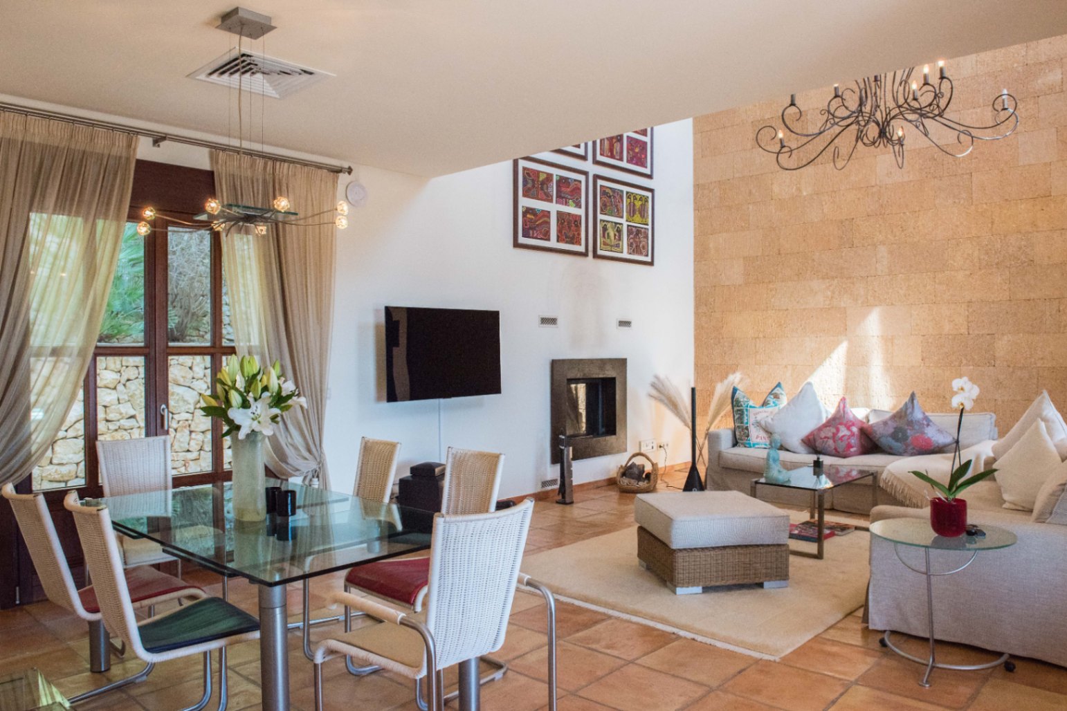 Open-plan living space featuring a textured stone accent wall, terracotta flooring, glass-top dining table with wicker chairs, and a lounge area with a fireplace.