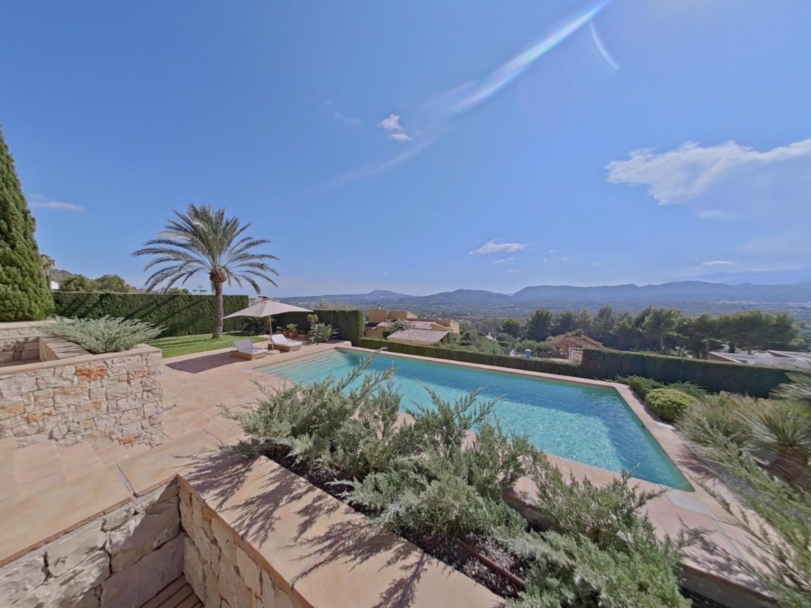 Outdoor swimming pool with a stone terrace and panoramic valley views. Features include sun loungers, a palm tree, and landscaped Mediterranean greenery.