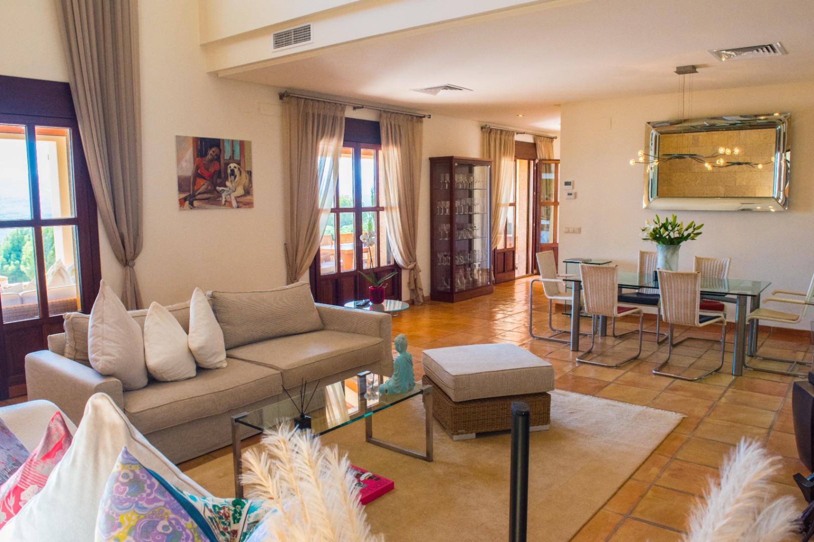 Spacious open-plan living area featuring terracotta tile floors, a glass-topped dining set, and a lounge area with views through multiple French doors.