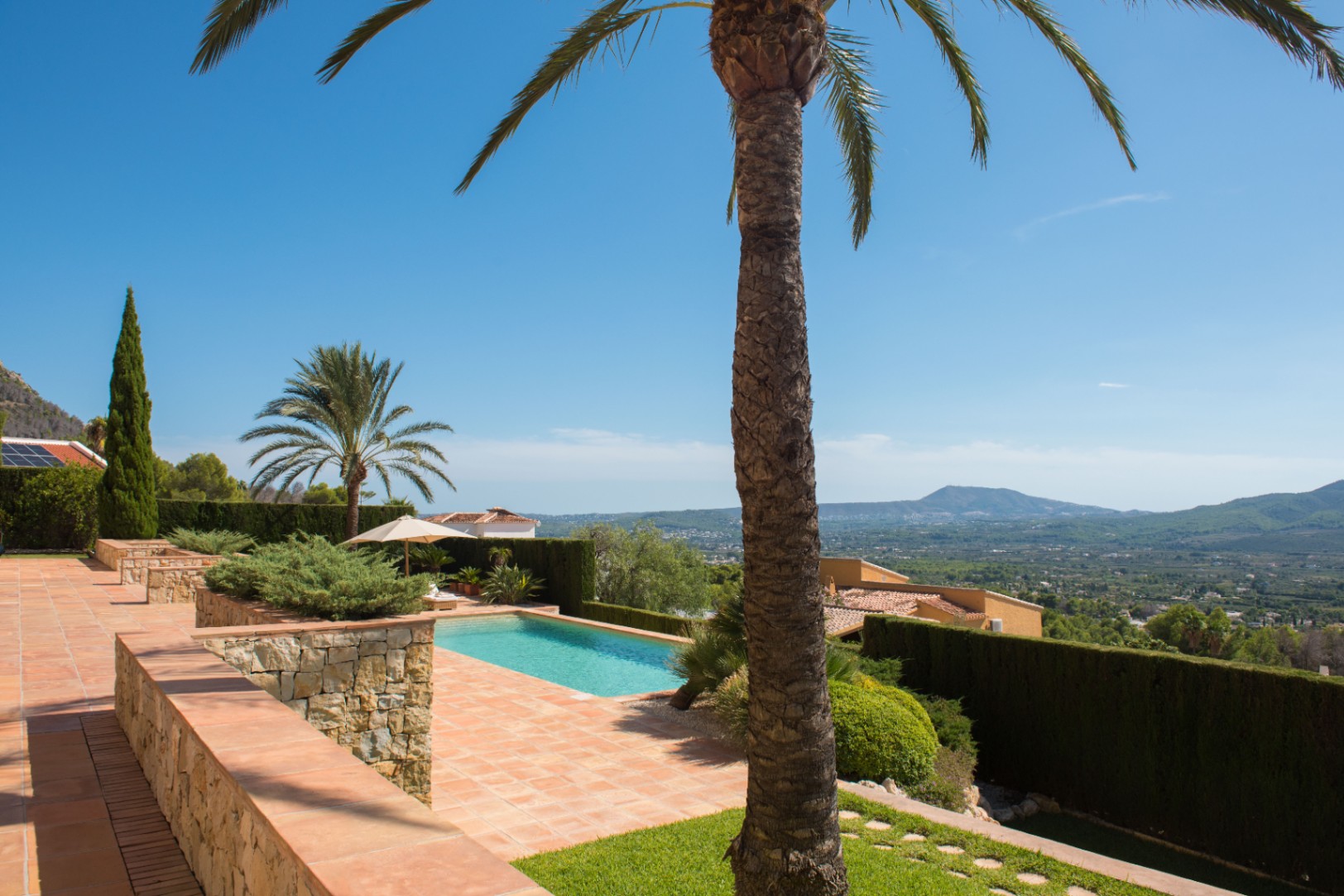 Terrace with terracotta tiles and stone walls featuring a swimming pool, palm trees, and panoramic views over a Mediterranean valley.