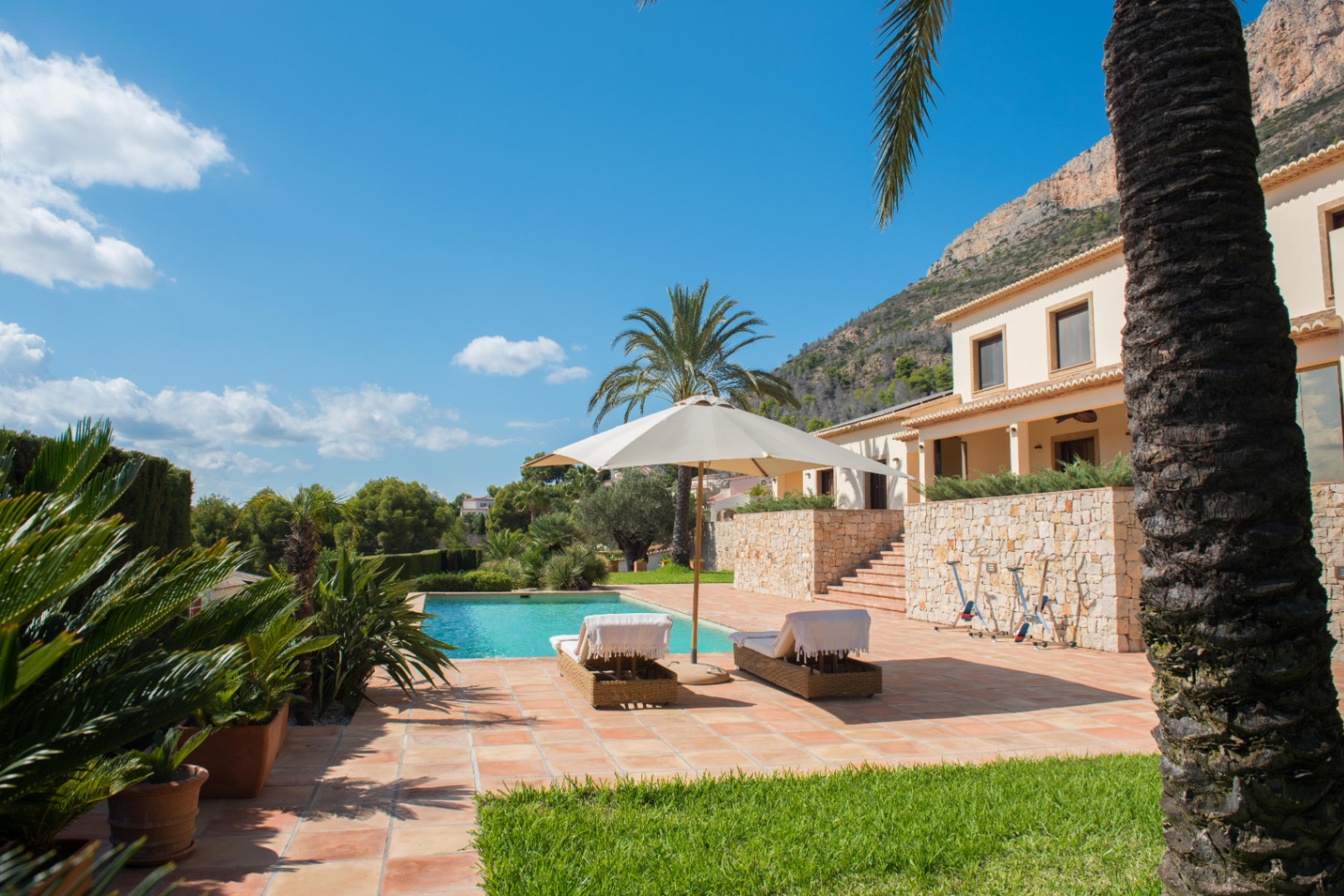 Terracotta terrace featuring a rectangular pool, wicker loungers, and dry stone walls. The landscaped garden includes palm trees set against a scenic mountain backdrop.