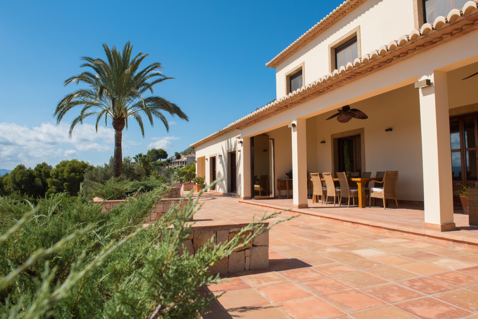 Terracotta tiled terrace featuring a covered dining area with wicker seating, overlooking a palm tree and lush Mediterranean greenery.