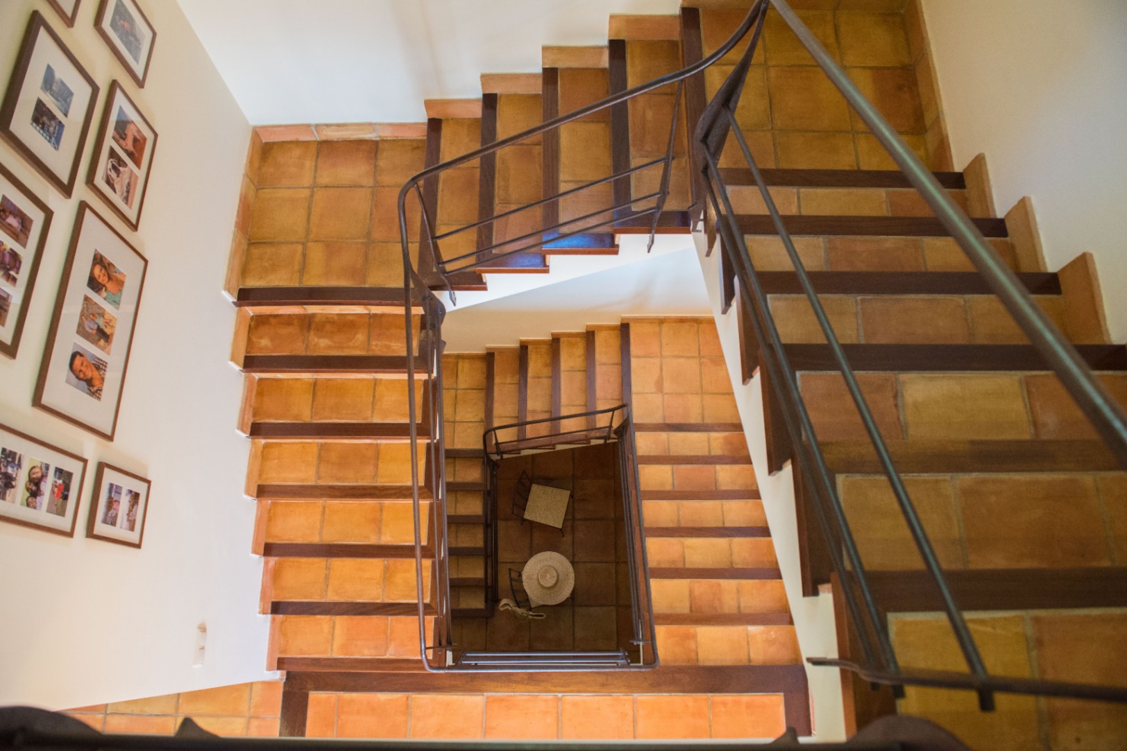 Top-down view of a multi-level staircase featuring terracotta tile treads with dark wood edging and a wrought iron handrail. Framed photographs line the adjacent white wall.