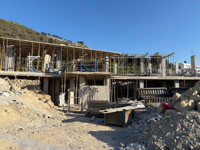 View of a multi-level villa under construction, showing reinforced concrete pillars, metal support props, and wooden formwork against a mountain backdrop.
