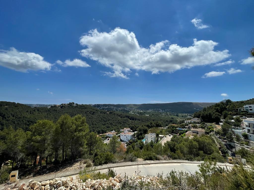 Elevated panoramic view overlooking a lush pine valley with scattered white villas, rolling hills, and a clear blue sky with light clouds.