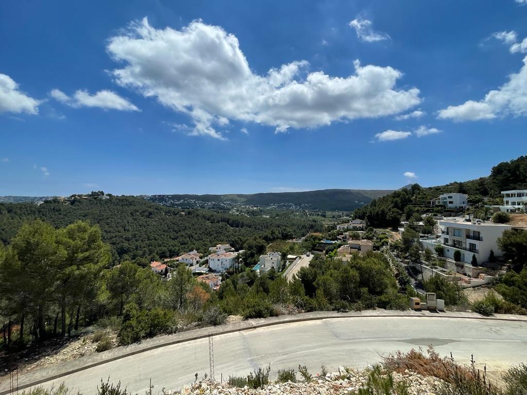 Panoramic elevated view of a Mediterranean valley featuring white villas nestled among pine-covered hills under a bright blue sky with scattered clouds.