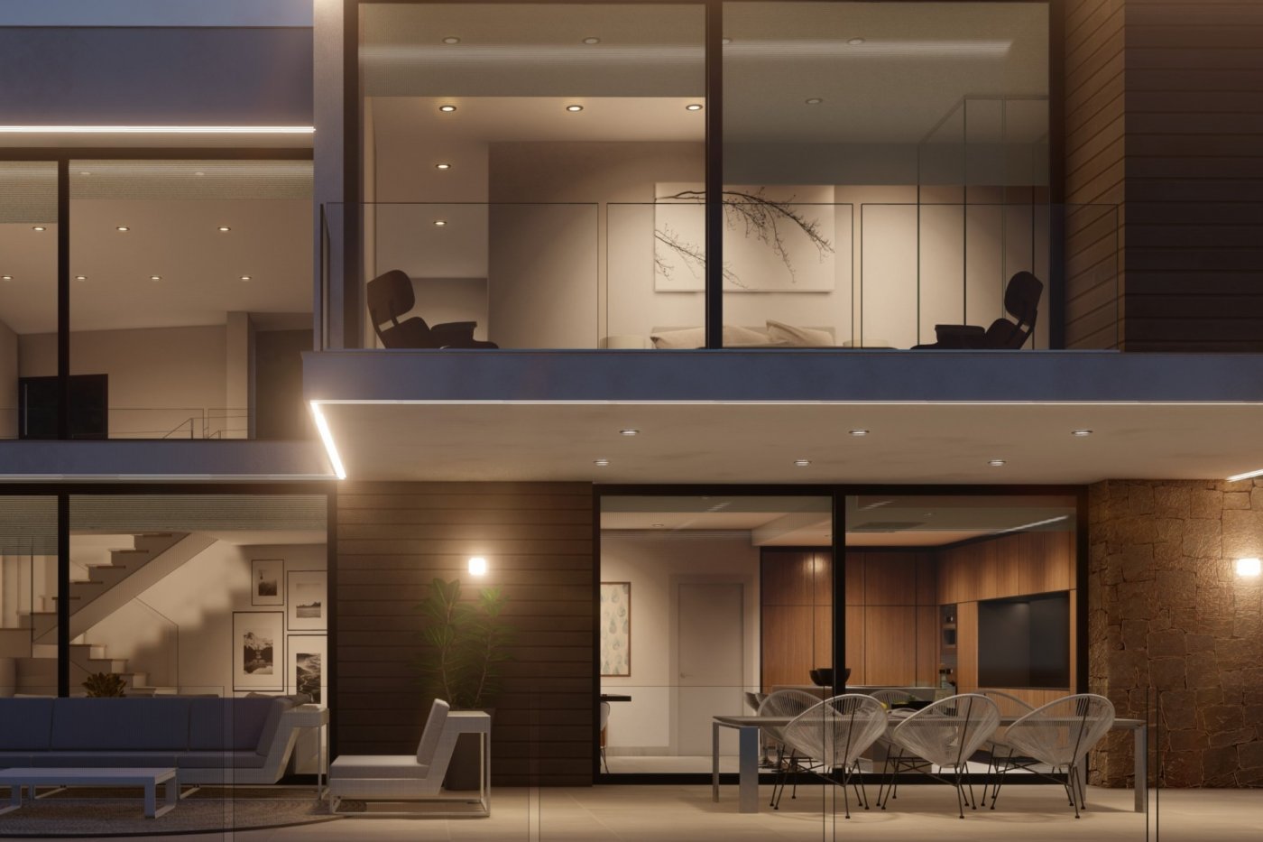 Exterior night view of a multi-level villa featuring a terrace with Acapulco chairs, stone masonry, and floor-to-ceiling windows illuminating the kitchen and upper balcony.
