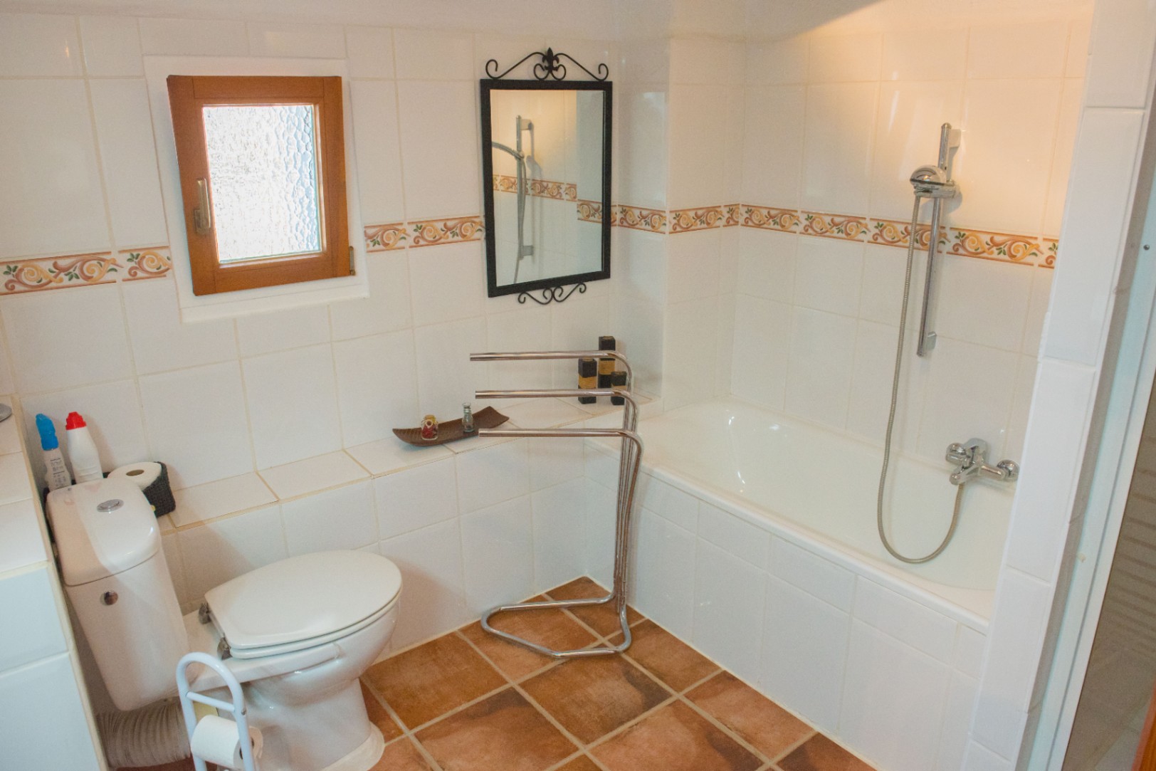 Bathroom featuring a built-in bathtub, terracotta floor tiles, and white wall tiling with decorative borders. Includes a wooden window and wrought-iron mirror.