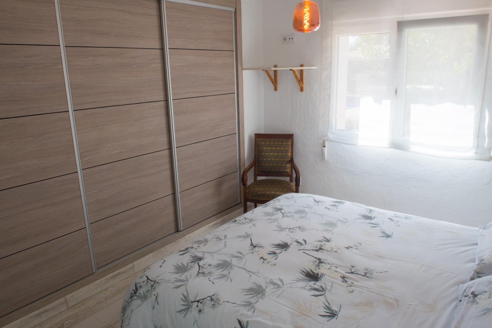 Bedroom featuring a large built-in wardrobe with sliding wood-effect panels, a vintage armchair, and natural light from a curtained window.