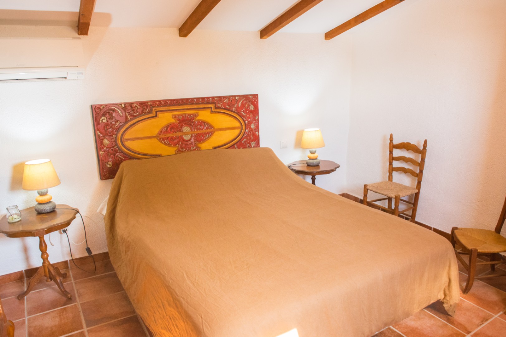 Bedroom featuring an ornate carved wooden headboard, exposed ceiling beams, terracotta tile flooring, and rustic wooden furniture.