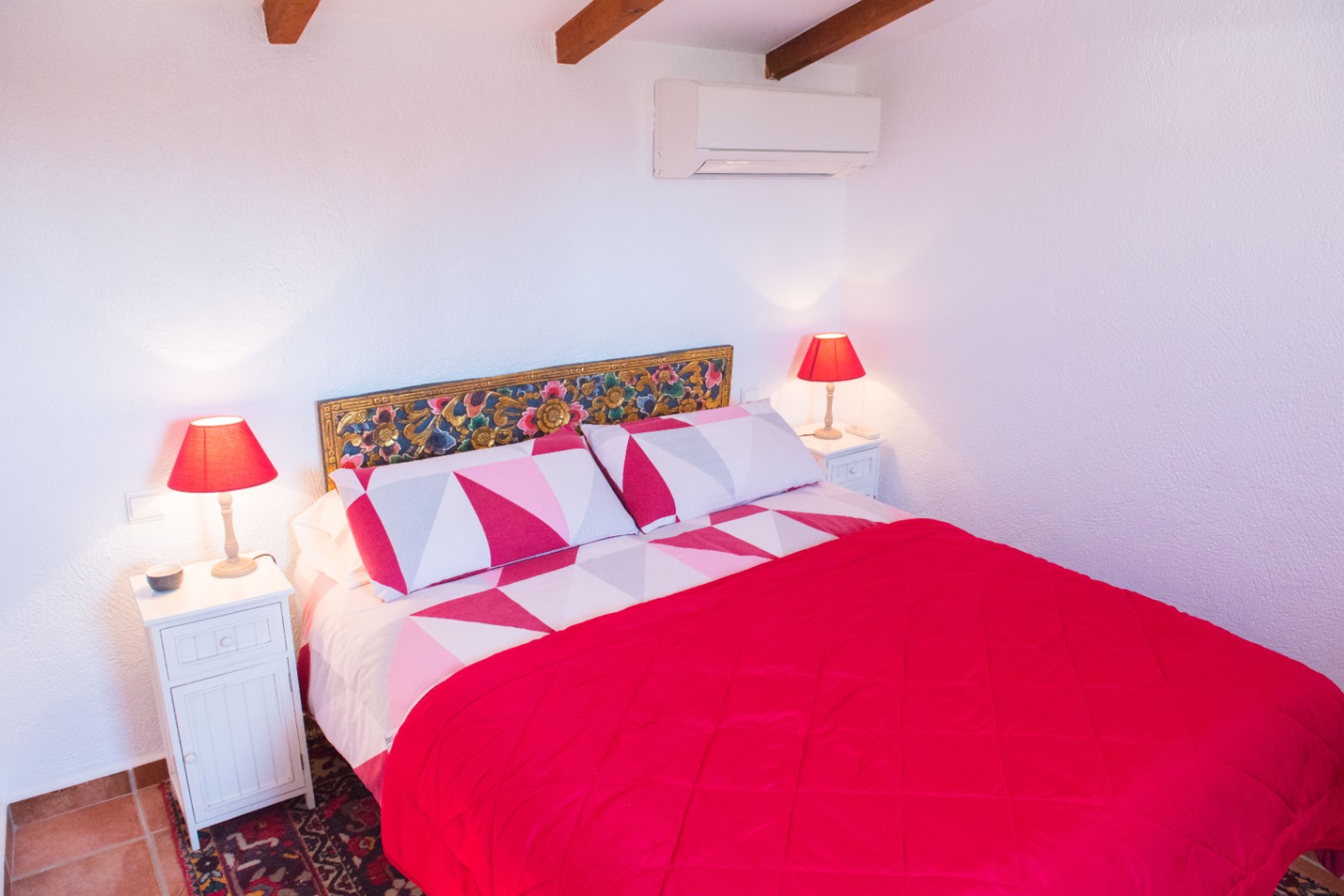Bedroom featuring exposed ceiling beams, an ornate carved headboard, and white nightstands with red lamps. Equipped with wall-mounted air conditioning.