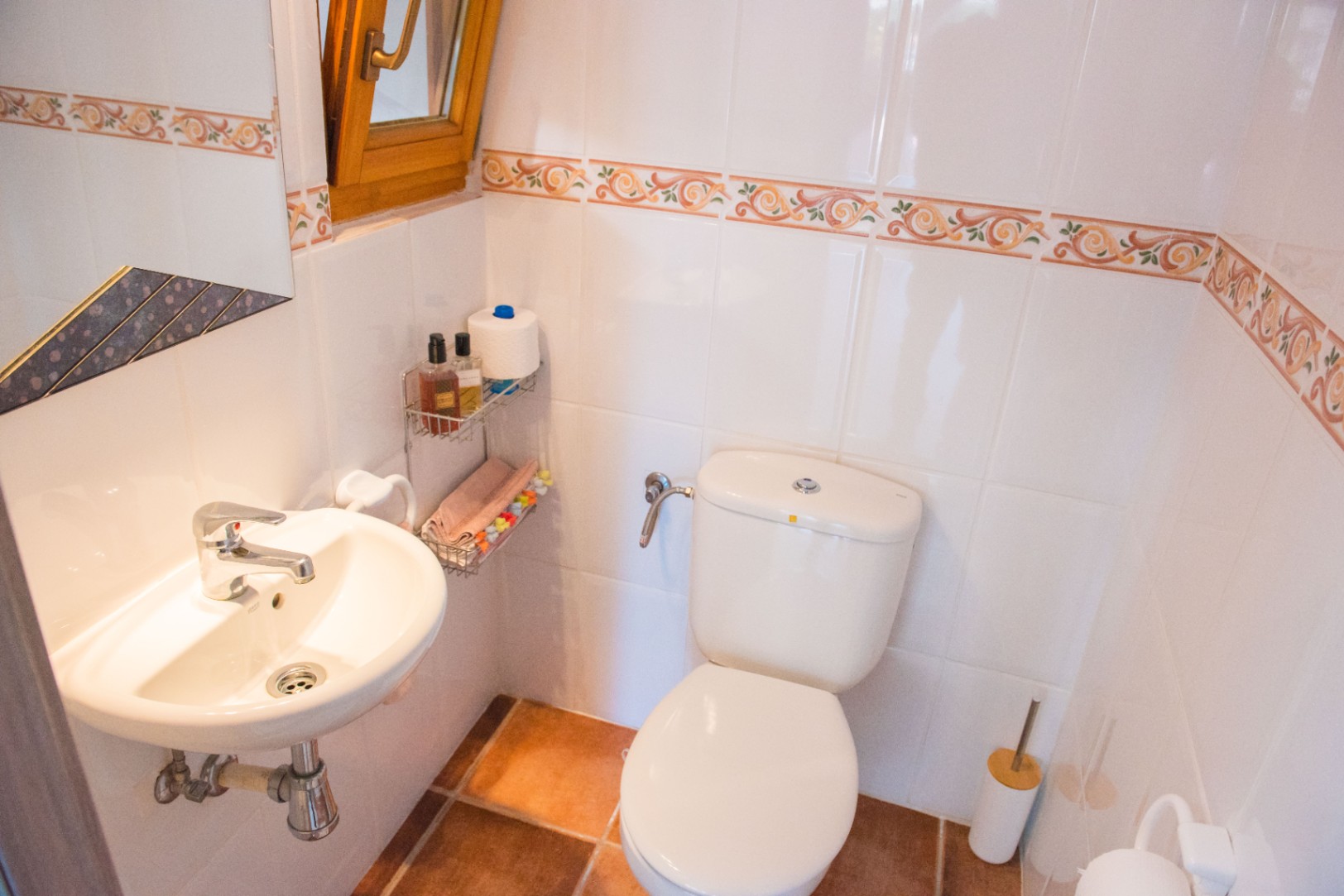 Compact bathroom featuring white wall tiles with decorative borders, a terracotta floor, wall-mounted ceramic sink, and a wooden-framed window for ventilation.