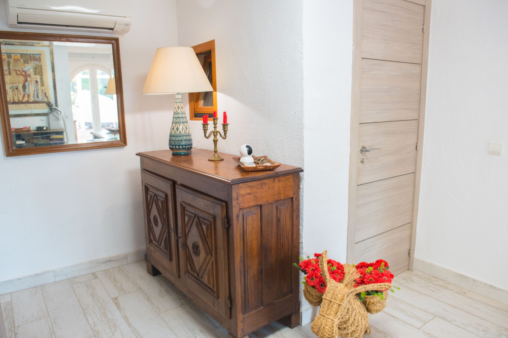 Entryway featuring a carved dark wood sideboard, a ceramic table lamp, and light wood-effect flooring. A mirror and air conditioning unit are mounted on the white textured wall.