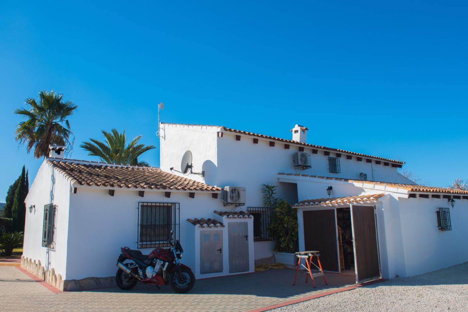 Exterior view of a white-washed villa featuring terracotta roof tiles, exposed wooden beams, and a paved driveway under a clear blue sky.
