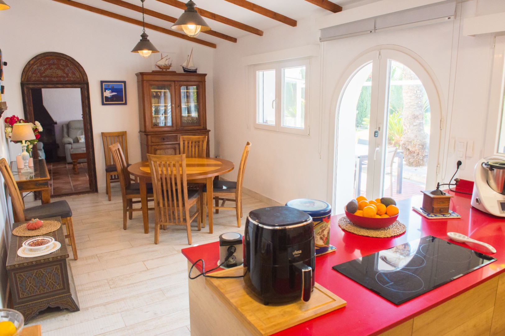 Kitchen featuring a red island with induction hob, wooden dining set, exposed ceiling beams, and arched glass doors to the patio.