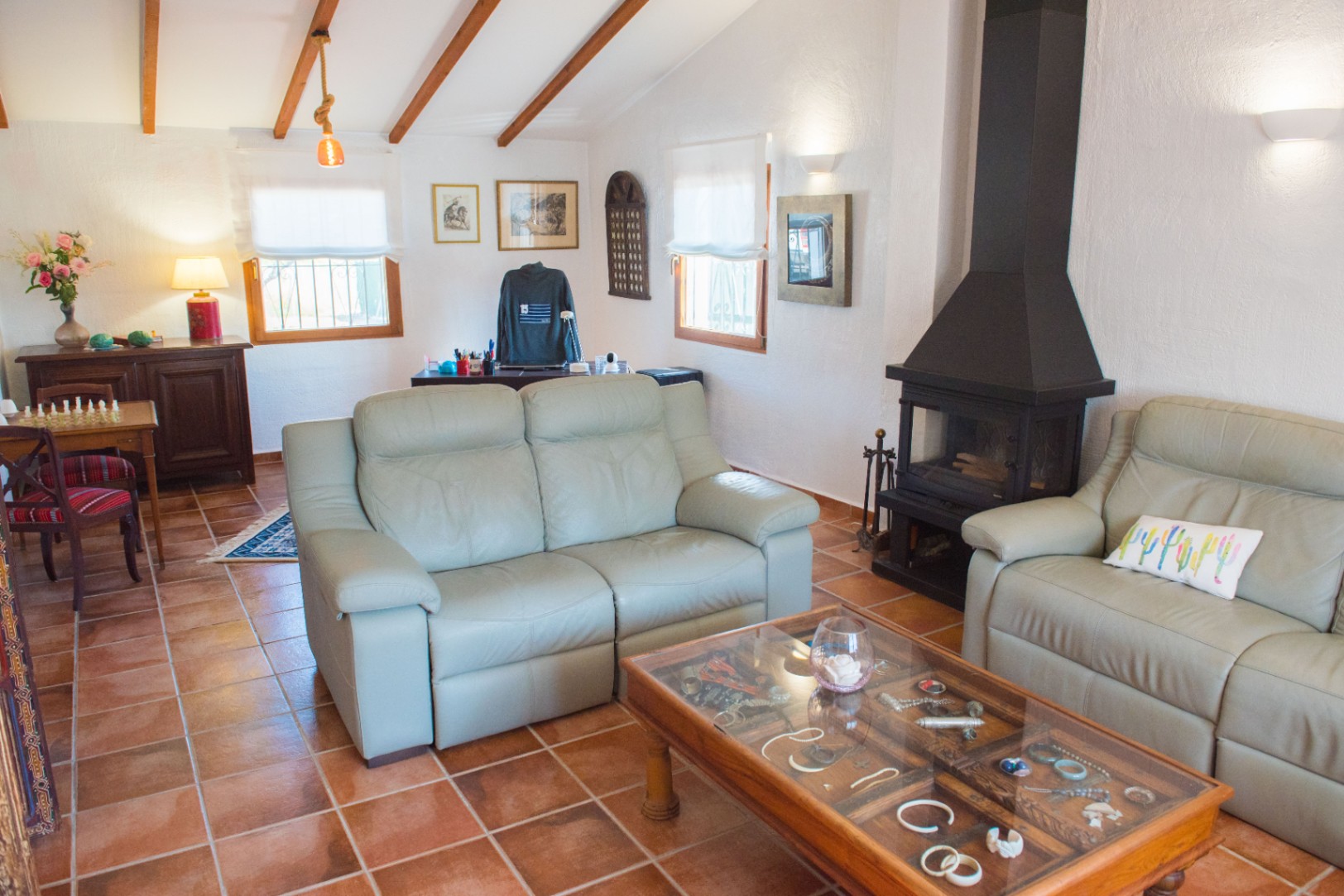 Living room featuring terracotta tile flooring, a black metal fireplace, and sloped ceilings with exposed wooden beams. Includes leather sofas and a glass-top display coffee table.