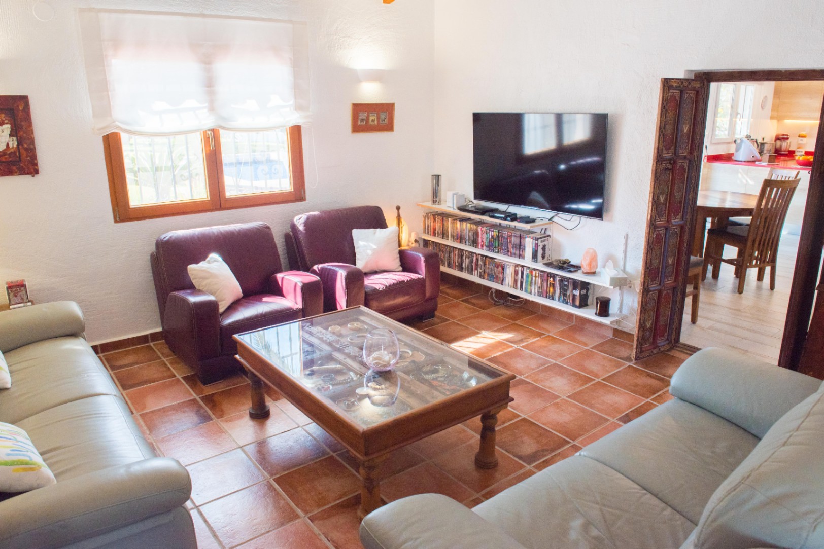 Living room with terracotta tile flooring, leather seating, and a glass-top coffee table. Features media shelving under a wall-mounted TV and a carved wooden door frame.