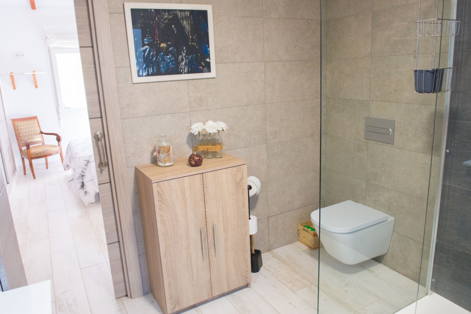 Modern bathroom featuring a wall-hung toilet, glass shower screen, and a light wood storage cabinet against neutral stone-effect wall tiles.