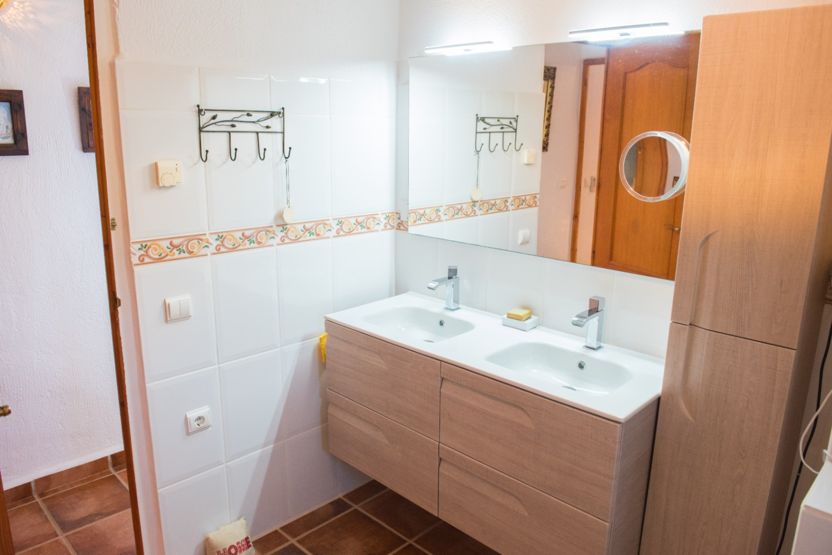 Modern double vanity in light wood with integrated white basins, chrome fixtures, and a large mirror featuring overhead lighting and a magnifying extension.