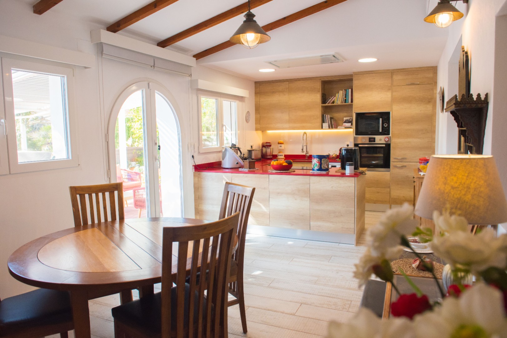Modern kitchen with light wood cabinetry, a bold red countertop, and integrated appliances, featuring a slanted ceiling with exposed beams and an adjacent dining area.