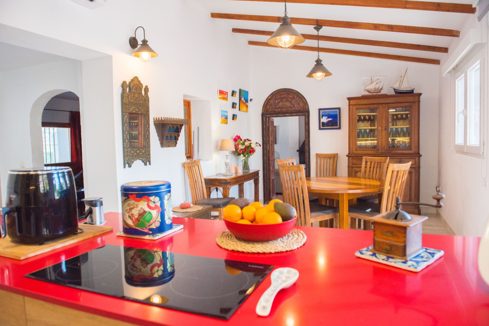 Open-plan kitchen featuring a bold red countertop and induction hob, overlooking a dining space with exposed ceiling beams and traditional wooden furniture.