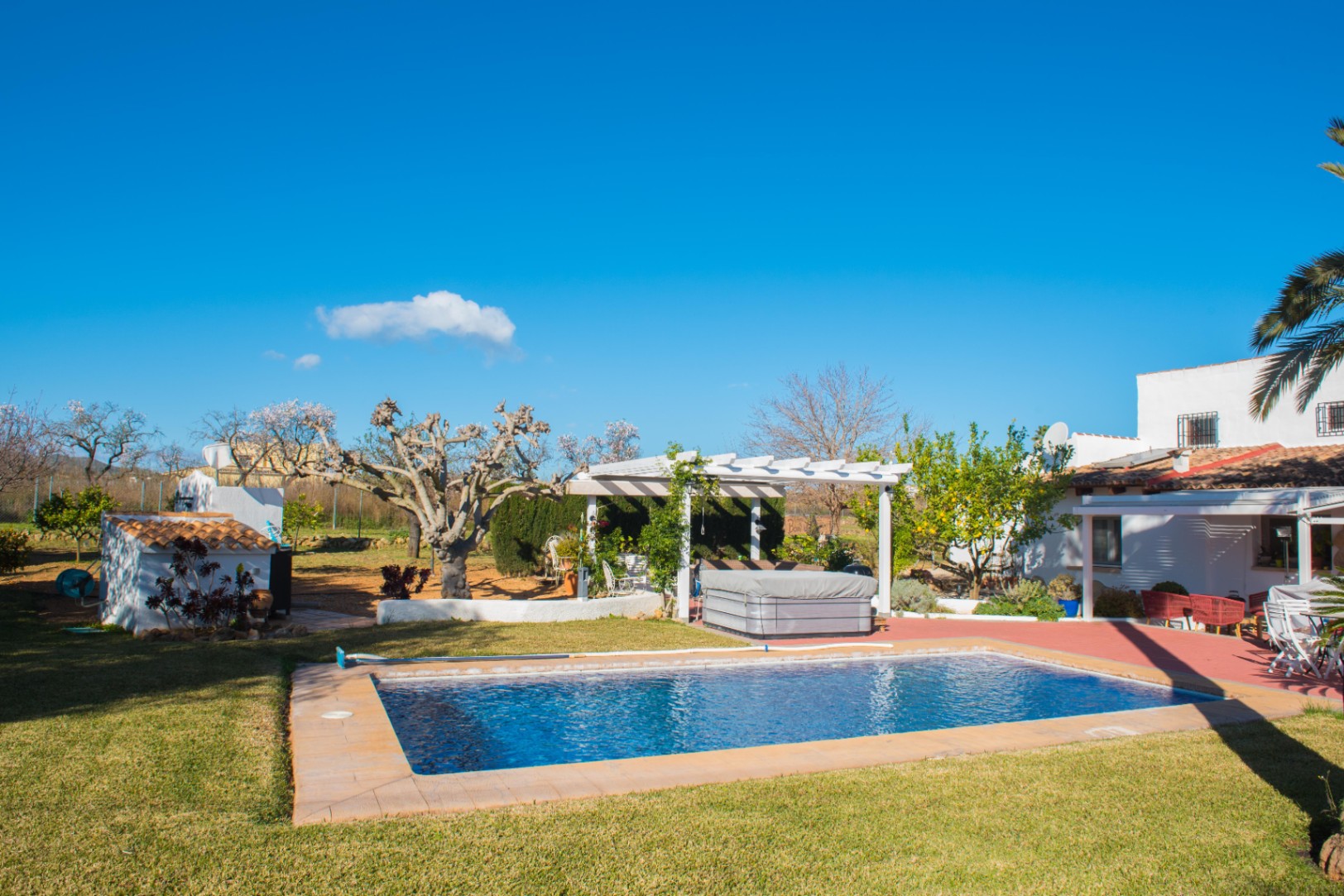 Outdoor area featuring a rectangular swimming pool, a manicured lawn, and a white pergola sheltering a hot tub, set against a Mediterranean-style villa.