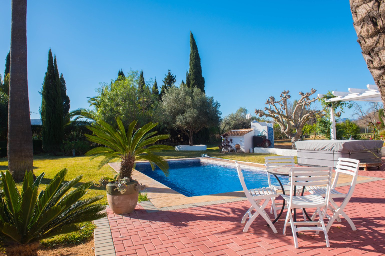 Outdoor terrace with red brick paving, featuring a rectangular swimming pool, white garden furniture, and a landscaped lawn with Mediterranean trees.