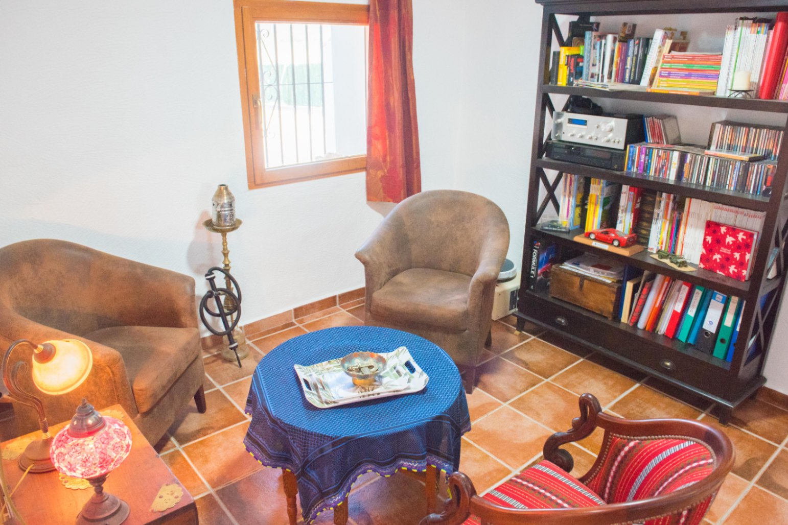 Reading area with terracotta tile flooring, a dark wood bookcase, and armchairs arranged around a round table covered with a blue cloth.